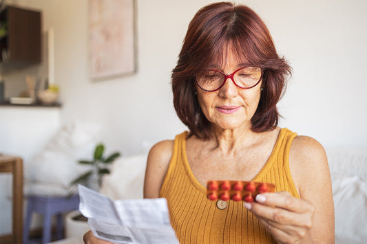 a mature woman wearing an ochre-colored top examines a package insert while holding a blister pack of hormone therapy pills