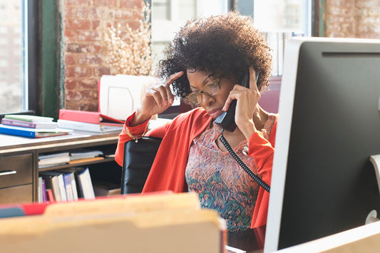 A woman with curly hair and glasses, wearing an orange cardigan, talks on the phone, discussing health insurance coverage for menopause-related care and treatments.