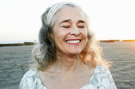Older woman with long gray hair standing on a beach at sunset, emphasizing her natural skin texture and signs of aging 