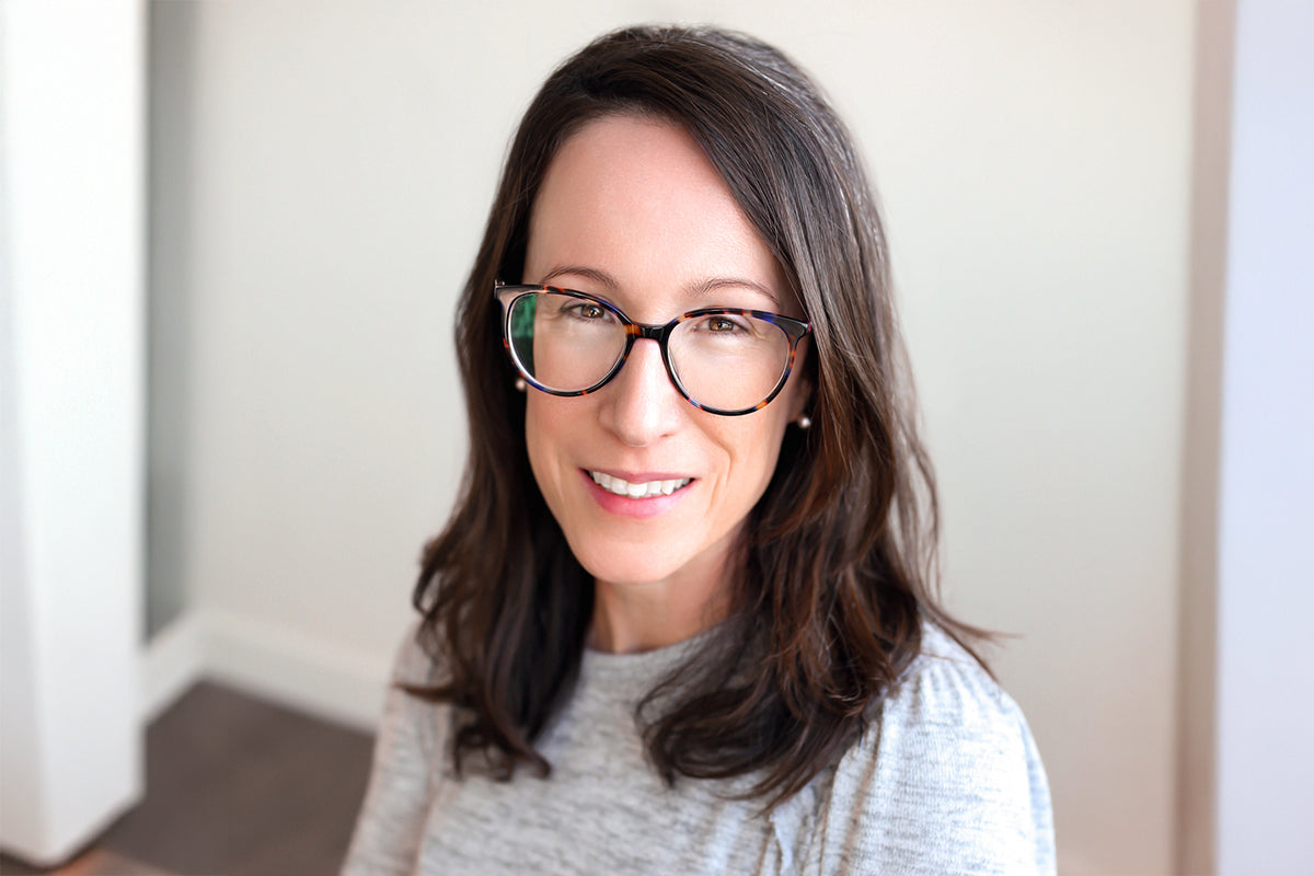 Portrait of Emily with long dark hair and glasses, wearing a light gray top, against a plain background