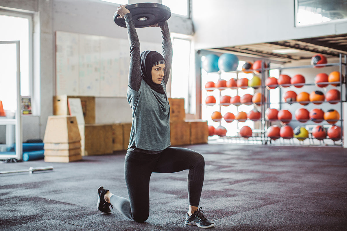 A woman wearing a hijab and workout gear performs a kneeling overhead press with a weight plate at a gym, demonstrating the kind of exercise that can build bone density and muscle mass during menopause.