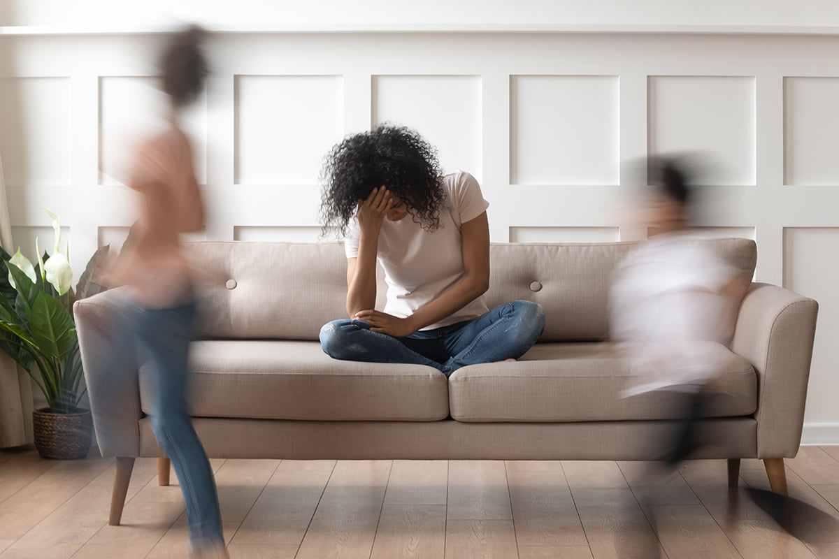 A woman sits on a beige couch with her head in her hand, looking overwhelmed, while two blurred children rush around her, capturing the chaos and emotional intensity often felt during menopause-related anger and irritability.