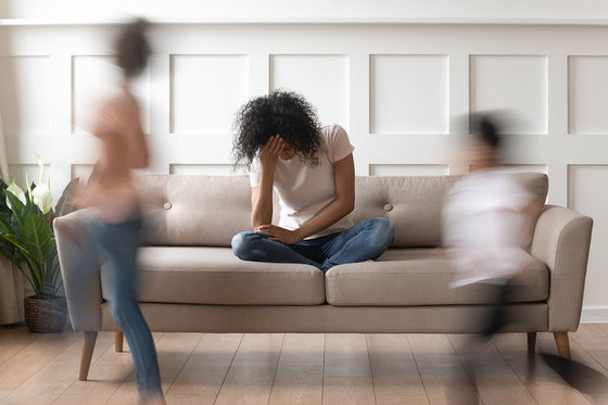 A woman sits on a beige couch with her head in her hand, looking overwhelmed, while two blurred children rush around her, capturing the chaos and emotional intensity often felt during menopause-related anger and irritability.