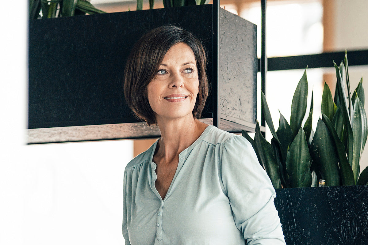 A woman stands confidently near some houseplants knowing she knows how to manage UTIs and urinary incontinence