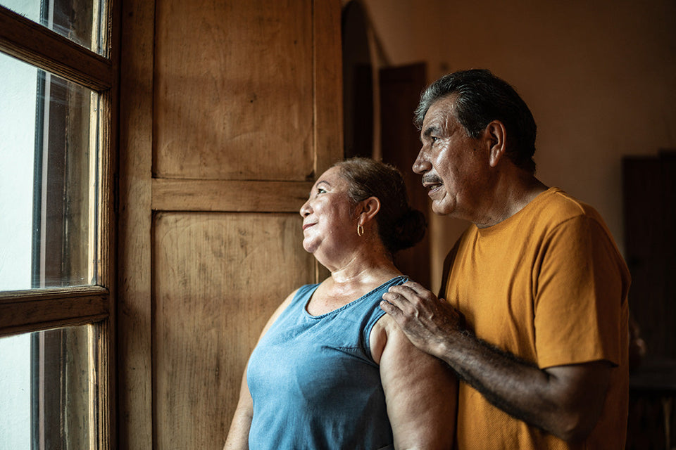 Middle-aged couple standing indoors near a window, the man gently resting his hand on the woman’s shoulder, conveying support and closeness in a warm setting.
