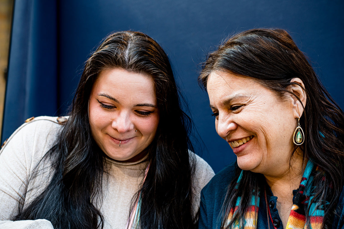 Close-up of a mother and daughter. The daughter, on the left, has long black hair and a lip piercing. The mother, on the right, is smiling, wearing a colorful scarf and teardrop earrings.