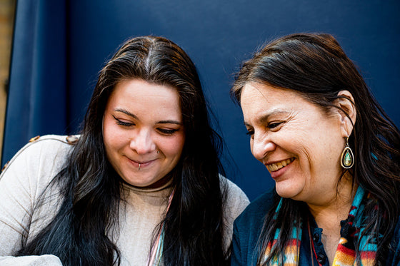 Close-up of a mother and daughter. The daughter, on the left, has long black hair and a lip piercing. The mother, on the right, is smiling, wearing a colorful scarf and teardrop earrings.