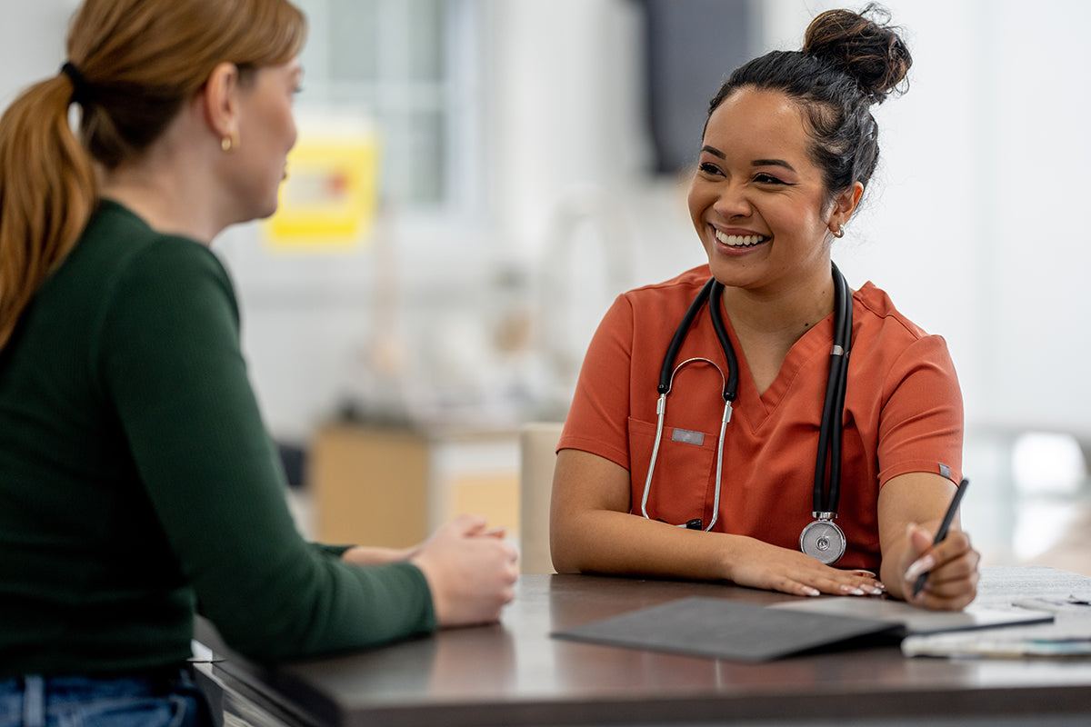 Middle-aged patient consulting with a healthcare professional in a clinic about recommended health screenings after age 40. The provider in orange scrubs points to a medical document, emphasizing preventive care and wellness.