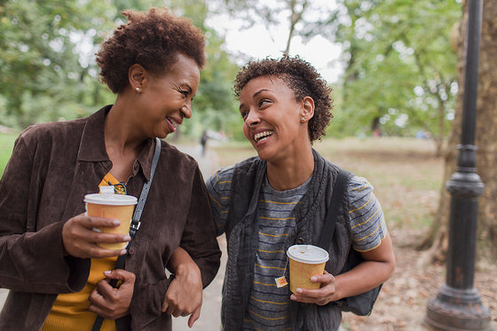Two smiling Black women, one older and one younger, walking together outdoors with coffee cups. They are looking at each other, suggesting a supportive relationship. POI can be challenging, and having a strong support system is important.
