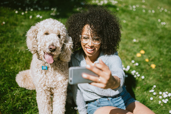 A woman with curly hair takes a selfie with her Goldendoodle dog in a grassy field.