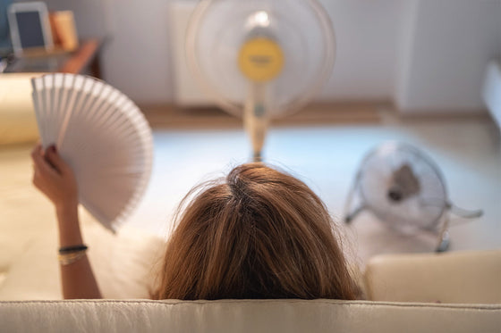 A woman lies on a couch, viewed from behind, as she tries to stay cool in the heat and from hot flashes. She holds a white hand fan, while two electric fans in the background spin to circulate air.  