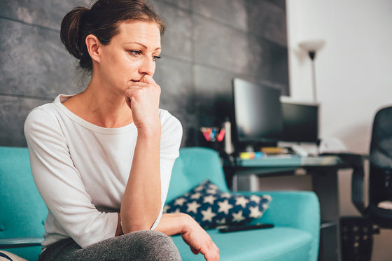 Middle-aged woman sitting pensively on a teal couch in a home office, reflecting stress or emotional discomfort—illustrating the experience of anxiety and irritability during menopause.